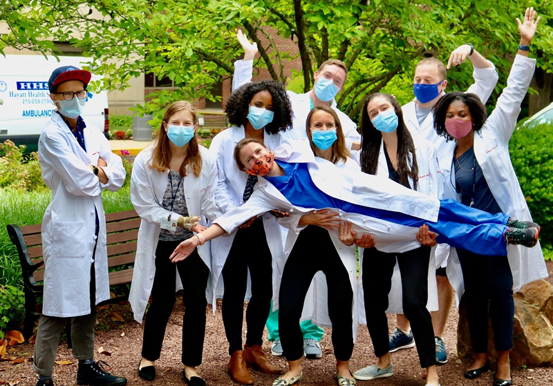 A group of nine medical residents in white coats and face masks pose for the camera outside among trees.